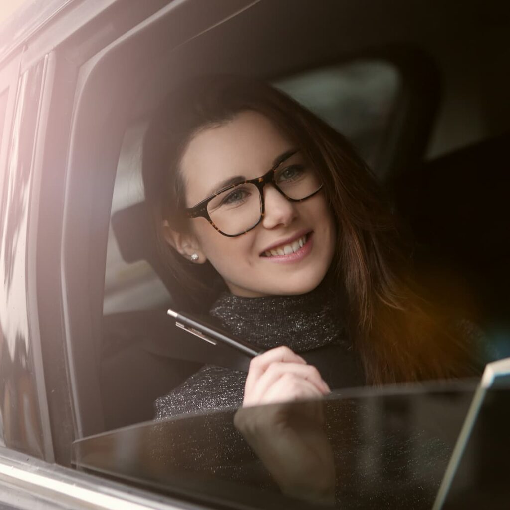 Eine zuversichtlich lächelnde, junge, dynamische und aufstrebende Frau sitzt im Auto auf dem Weg zu ihrem nächsten Geschäftstermin. Sie hält einen Stift und ein Buch in der Hand und freut sich voller Erwartung.