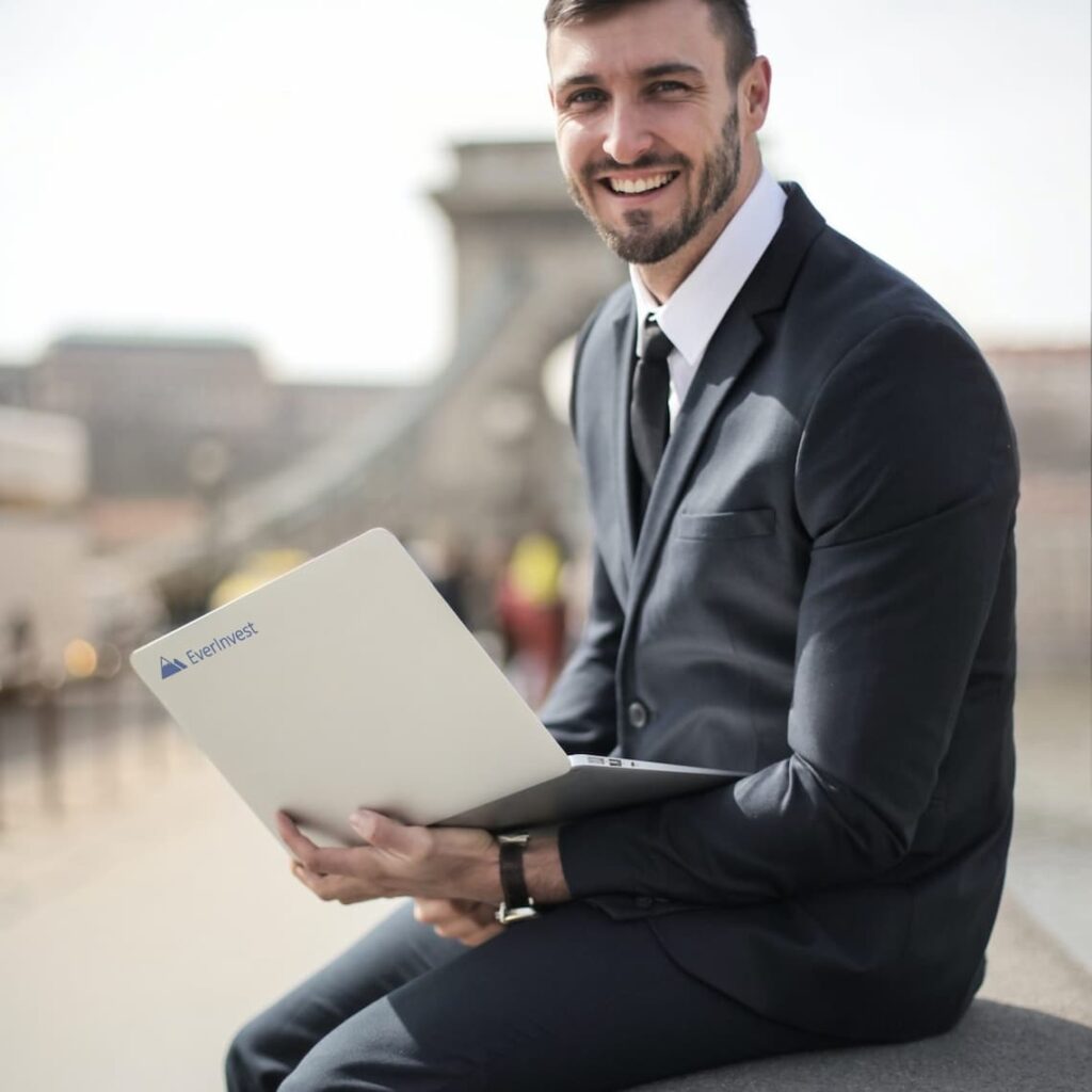 Unser Finanzprofi für Risikomanagement sitzt auf einer Bank mit einem Laptop auf seinem Schoß und lächelt freundlich aufgeschlossen. Auf dem Laptop ist das EverInvest Logo zu sehen welches alle unsere Kursleiter haben.